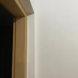 Gładka, biała ściana po malowaniu z widoczną ościeżnicą drzwiową z jasnego drewna. Fragment wykończonego wnętrza, minimalistyczny styl.