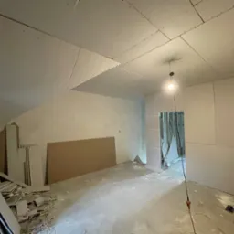 Pokój w trakcie remontu: ściany i sufit pokryte płytami kartonowo-gipsowymi, widoczne łączenia. Podłoga posprzątana, przygotowana do dalszych prac wykończeniowych.