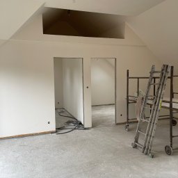 Lukasz Myszak - Świeżo pomalowane, jasne wnętrze poddasza ze schodami i rusztowaniem. Widoczne kable elektryczne i surowa podłoga. Dwa otwory drzwiowe prowadzą do kolejnych pomieszczeń.