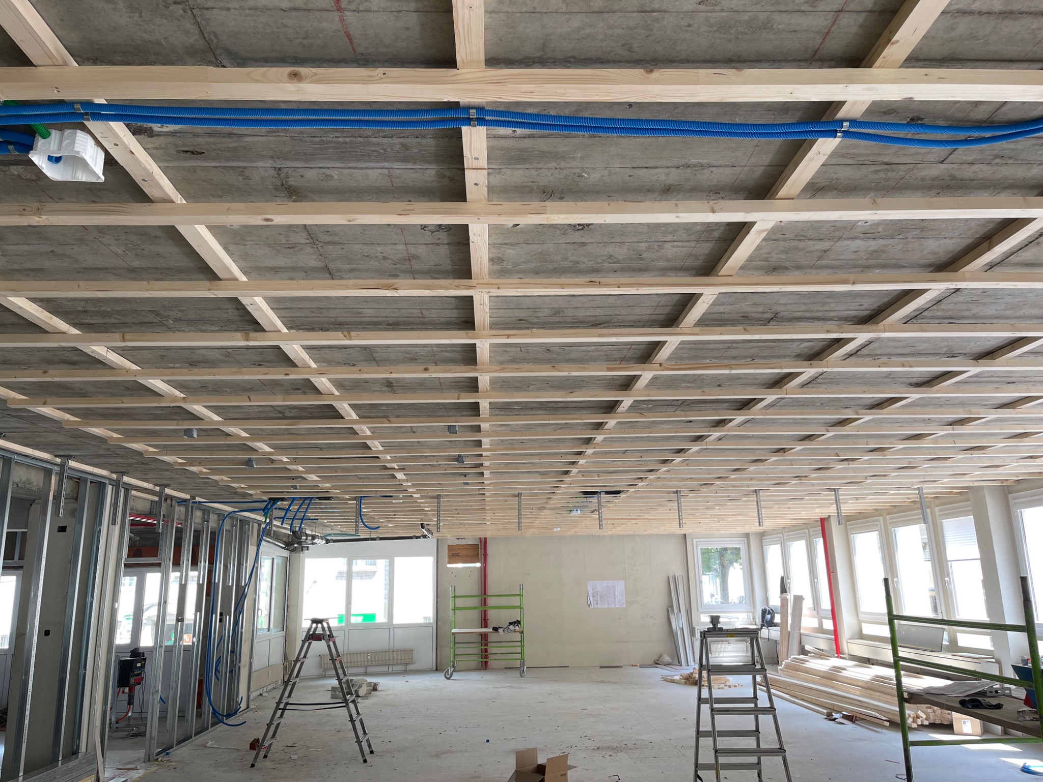 Sufit podwieszany z drewnianą konstrukcją i niebieskimi rurami instalacji w surowym wnętrzu. Metalowe profile ścian działowych w tle.