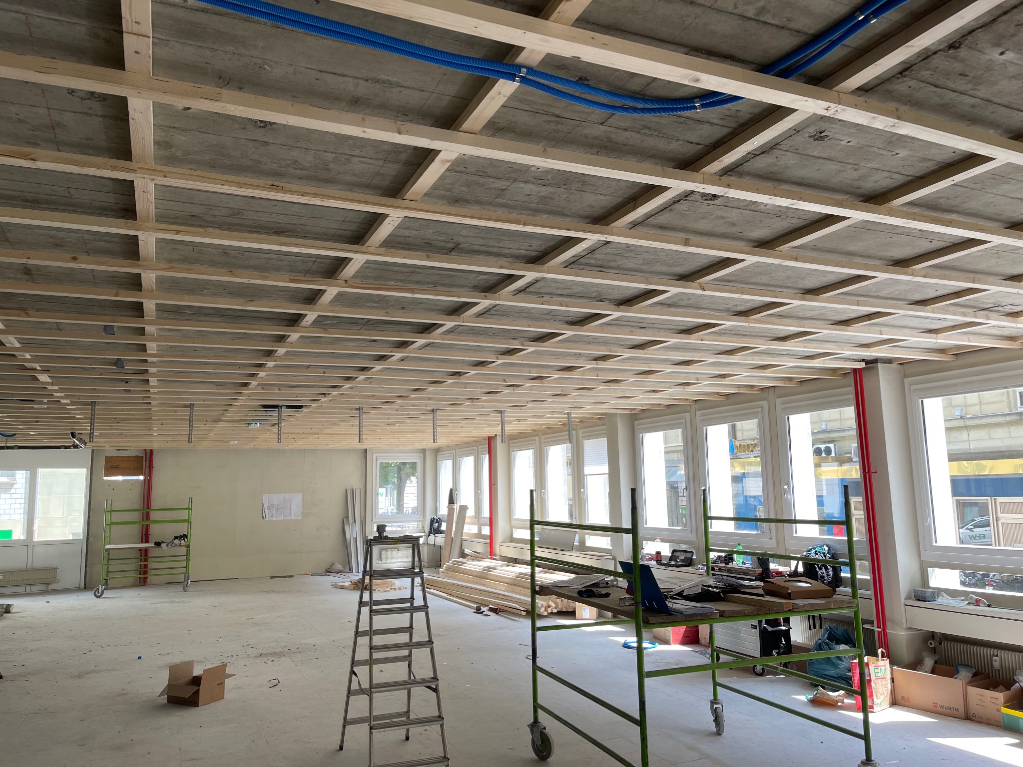 Montaż drewnianej konstrukcji sufitu podwieszanego w jasnym wnętrzu z licznymi oknami. Widoczne narzędzia i materiały budowlane.