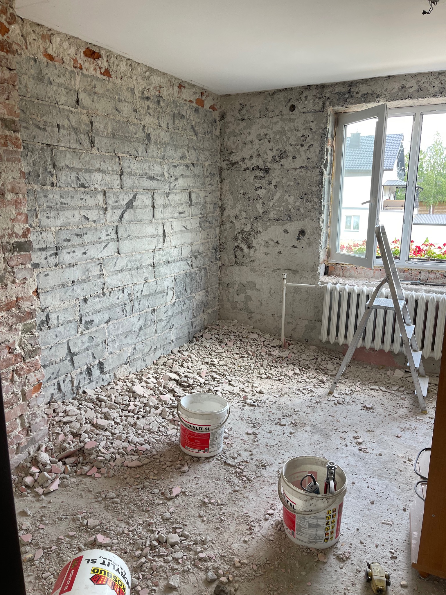 Pokój w trakcie skuwania tynku ze ścian, widoczne fragmenty cegieł, gruz na podłodze, drabina przy oknie i wiaderka z materiałami budowlanymi.