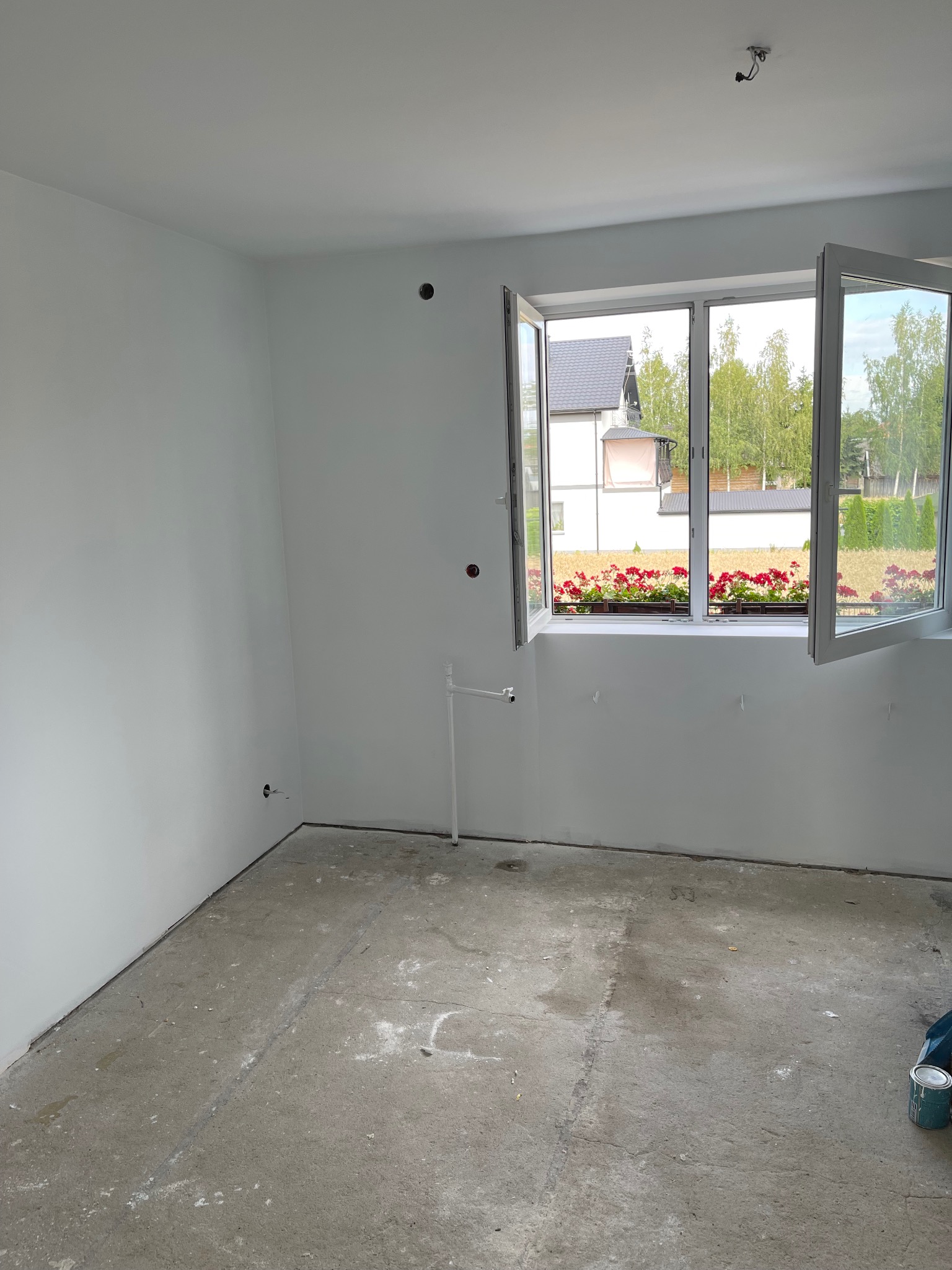 Białe ściany w pokoju po malowaniu, widoczne ślady farby na podłodze, okno z widokiem na zieleń i czerwone kwiaty. Wnętrze w trakcie remontu, przygotowane do dalszych prac.