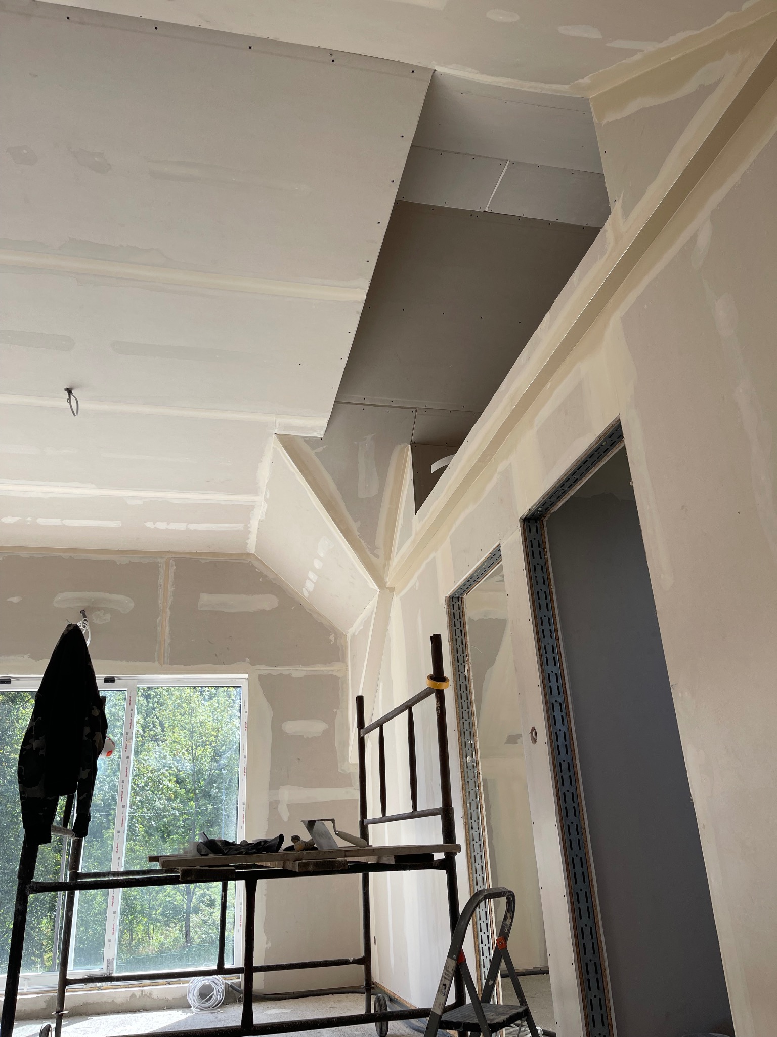 Wnętrze w trakcie wykańczania: ściany i sufit z płyt kartonowo-gipsowych, szpachlowane łączenia, metalowe rusztowanie z narzędziami, widok na okno i drzewa.
