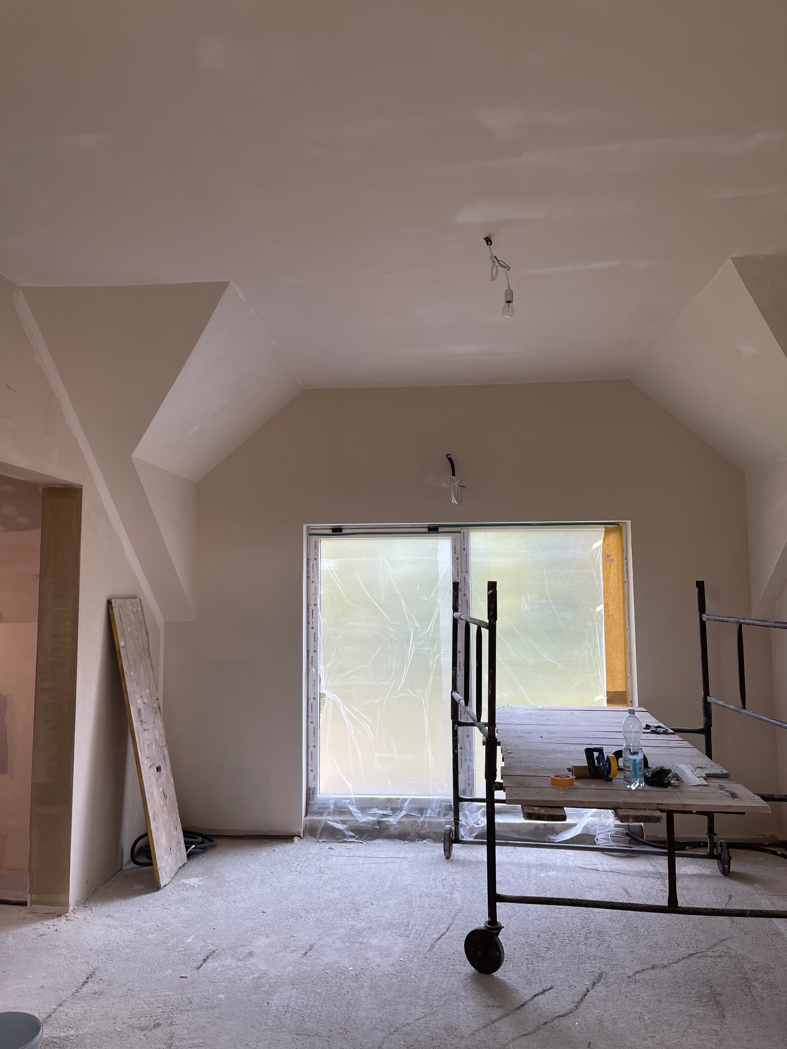 Pokój na poddaszu po szpachlowaniu, z przygotowaniem do malowania. Okno zabezpieczone folią, widoczne rusztowanie i narzędzia. Sufit ze skosami, surowy stan wykończenia.