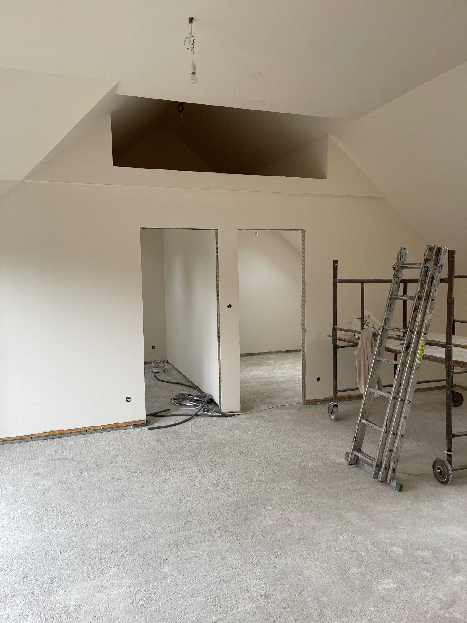 Świeżo pomalowane, jasne wnętrze poddasza ze schodami i rusztowaniem. Widoczne kable elektryczne i surowa podłoga. Dwa otwory drzwiowe prowadzą do kolejnych pomieszczeń.