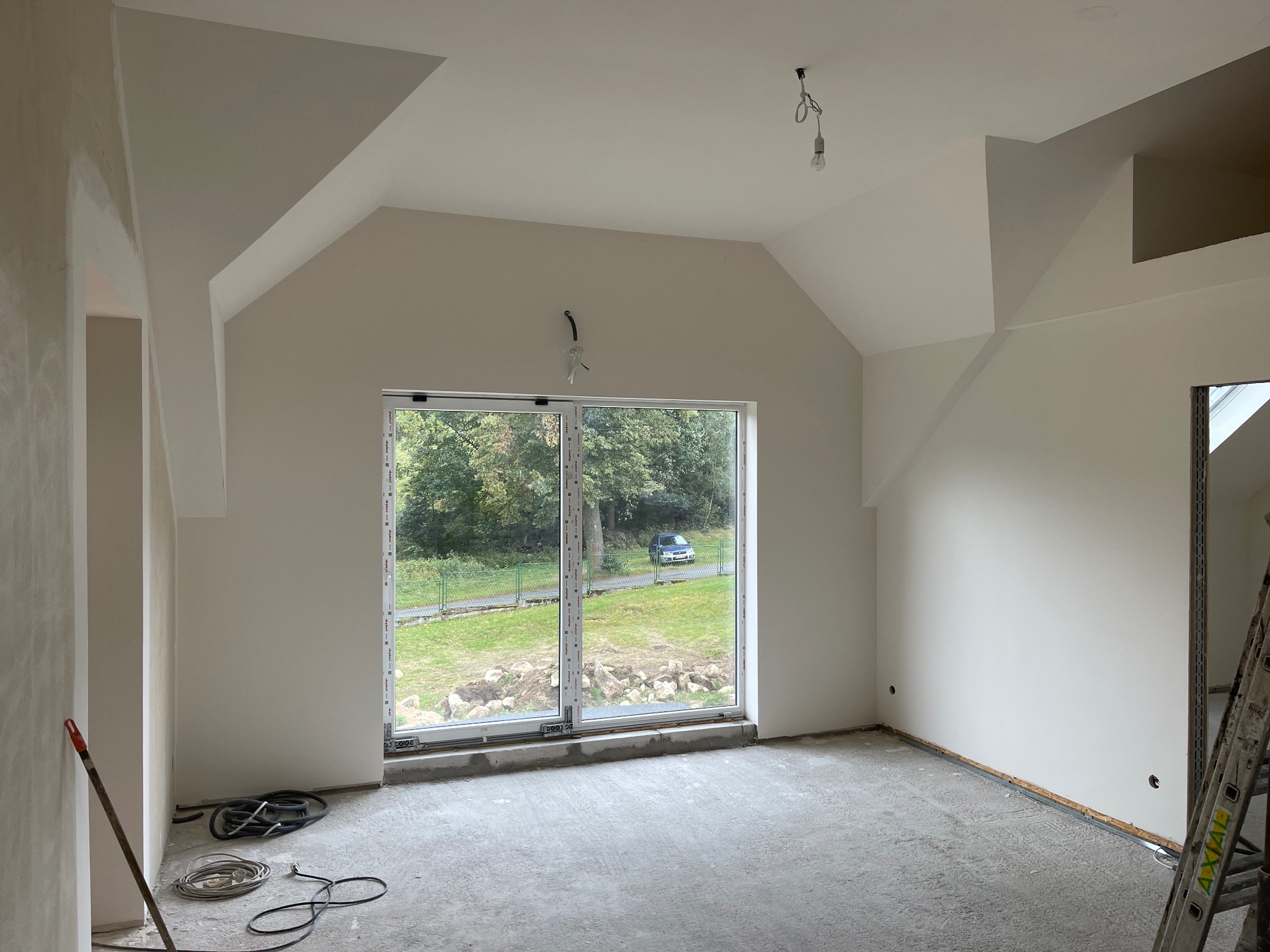 Pokój po gładzi i malowaniu na poddaszu z oknem na ogród. Widoczne kable elektryczne, drabina i narzędzia, gotowy do dalszych prac wykończeniowych.