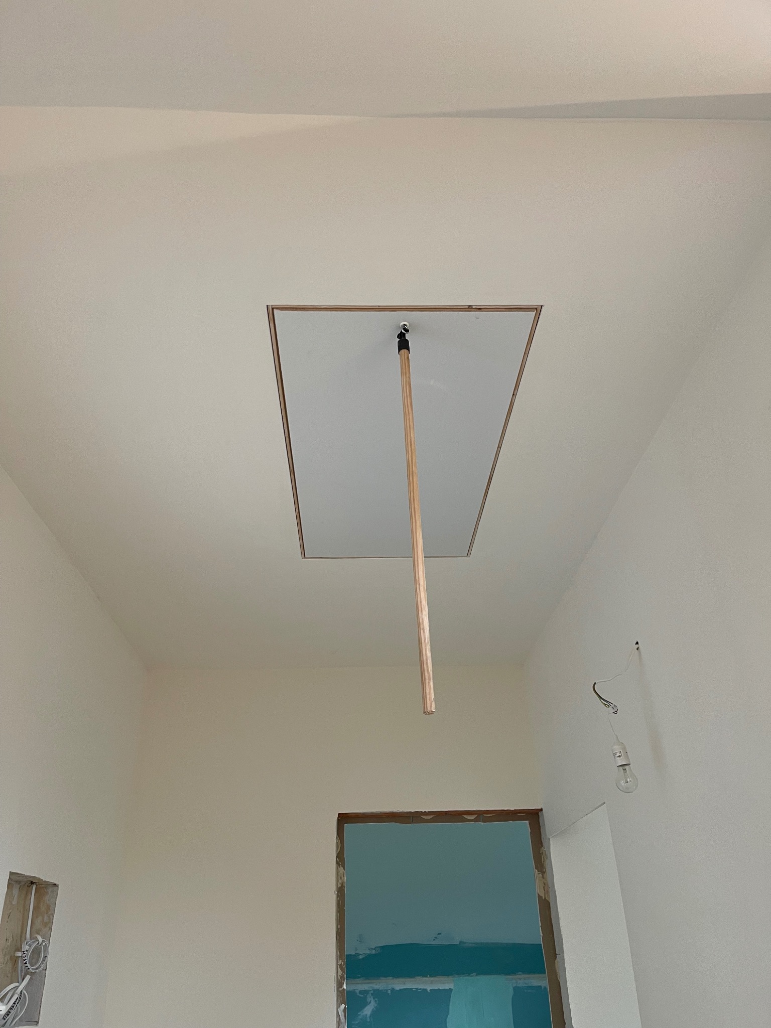 Biały sufit z klapą inspekcyjną, drewniany drążek do otwierania, ściany po malowaniu na jasny kolor, widoczne niedokończone prace.