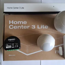Zestaw Fibaro Home Center 3 Lite: centrala, czujniki ruchu i otwarcia. Elementy systemu inteligentnego domu na kartonowym pudełku. Prezentacja komponentów.