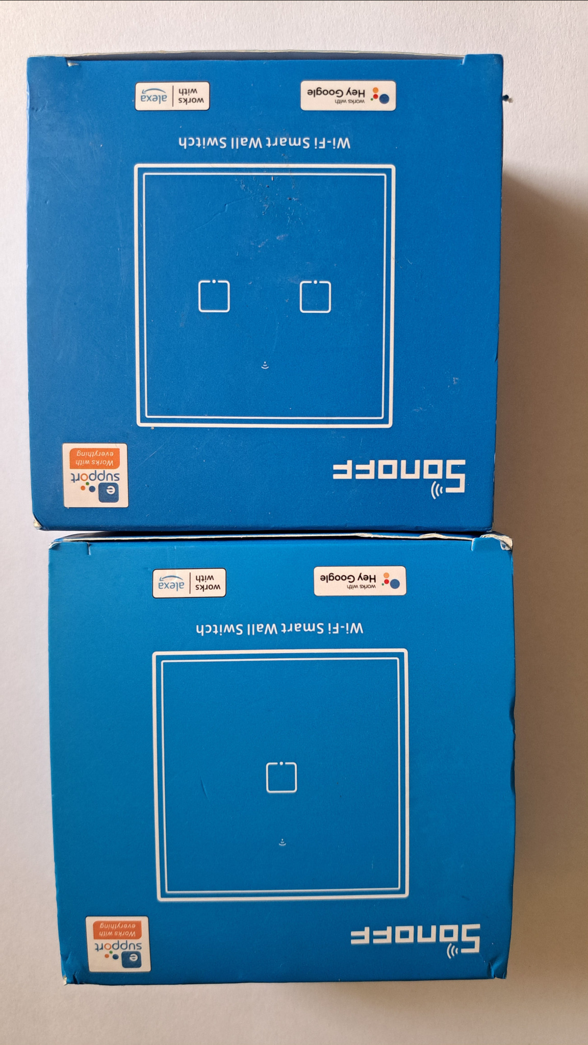 Dwa opakowania niebieskich, inteligentnych włączników ściennych Wi-Fi Sonoff. Opakowania wskazują na kompatybilność z Alexa i Google Home.