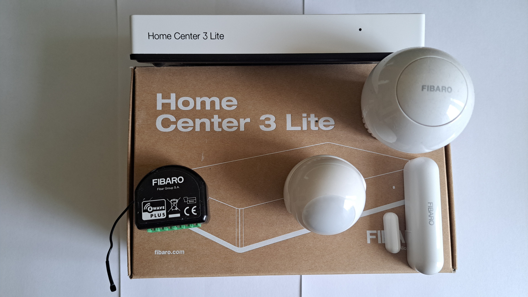 Zestaw Fibaro Home Center 3 Lite: centrala, czujniki ruchu i otwarcia. Elementy systemu inteligentnego domu na kartonowym pudełku. Prezentacja komponentów.