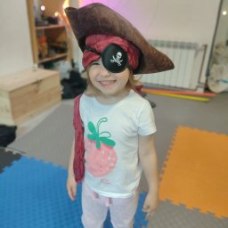 Dzikie Animacje - Uśmiechnięta dziewczynka w stroju pirata z opaską na oku z motywem czaszki i kapeluszu. Kolorowe puzzle piankowe na podłodze, tęczowe oświetlenie w tle.