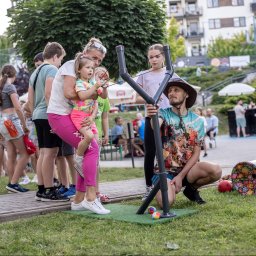 Dzikie Animacje - Impreza plenerowa w Przemyślu: animator z dziećmi celują z dużej procy do celu. W tle widoczni inni uczestnicy zabawy i budynki mieszkalne.