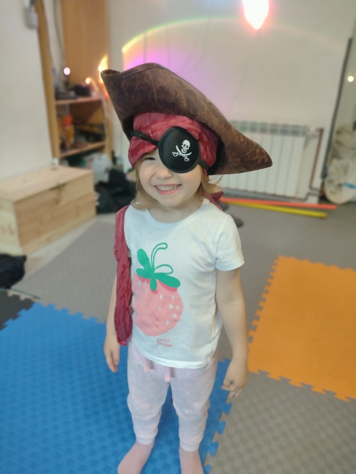 Uśmiechnięta dziewczynka w stroju pirata z opaską na oku z motywem czaszki i kapeluszu. Kolorowe puzzle piankowe na podłodze, tęczowe oświetlenie w tle.