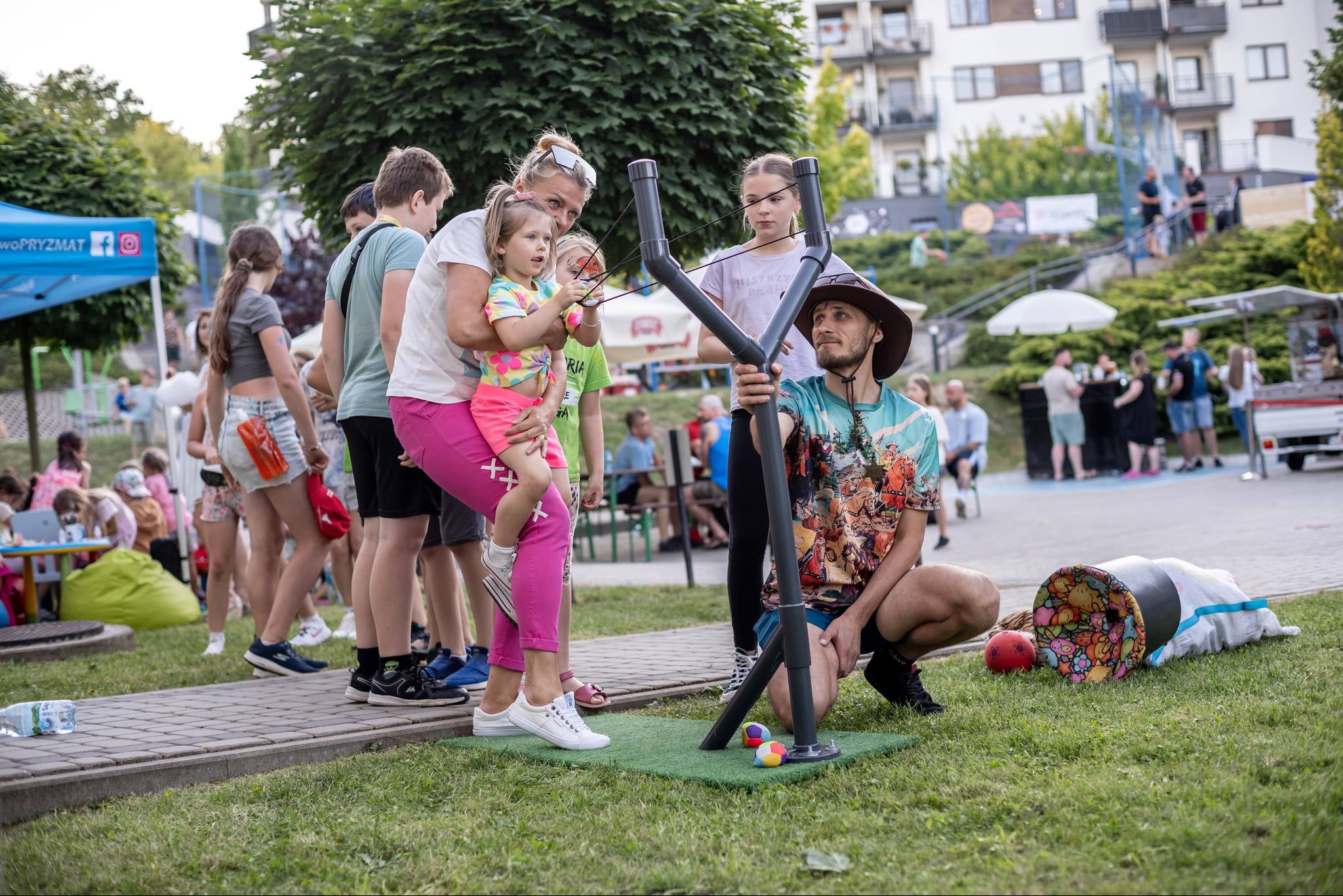 Impreza plenerowa w Przemyślu: animator z dziećmi celują z dużej procy do celu. W tle widoczni inni uczestnicy zabawy i budynki mieszkalne.