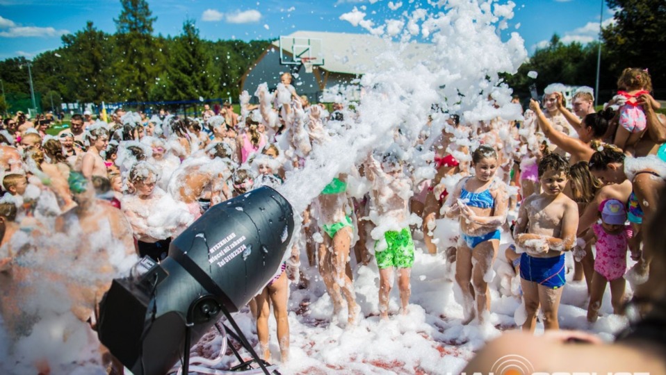 Piana party w plenerze: tłum dzieci i dorosłych w pianie, z dyszy leci strumień piany. W tle budynek z koszem do koszykówki i drzewa. Słoneczny dzień.
