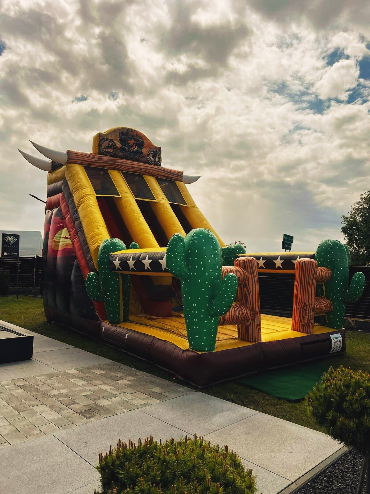 Tematyczny dmuchany zamek w stylu kowbojskim z motywem kaktusów i byka, ustawiony na zewnątrz na trawie, pod pochmurnym niebem, częściowo widoczny chodnik.