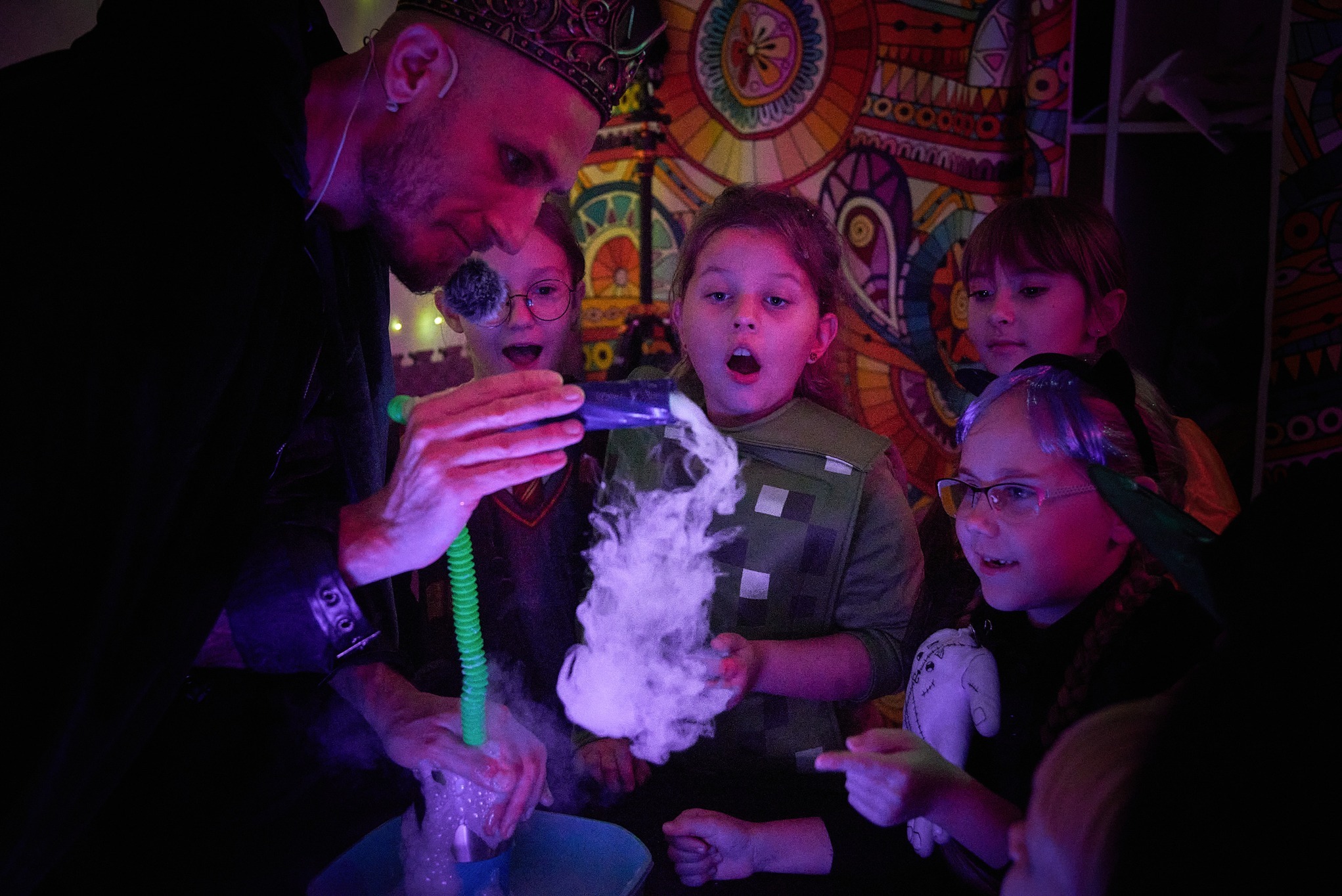Animator w koronie prezentuje dym z rekwizytu dzieciom podczas pokazu na tle kolorowej dekoracji. Dzieci z zaciekawieniem obserwują pokaz.
