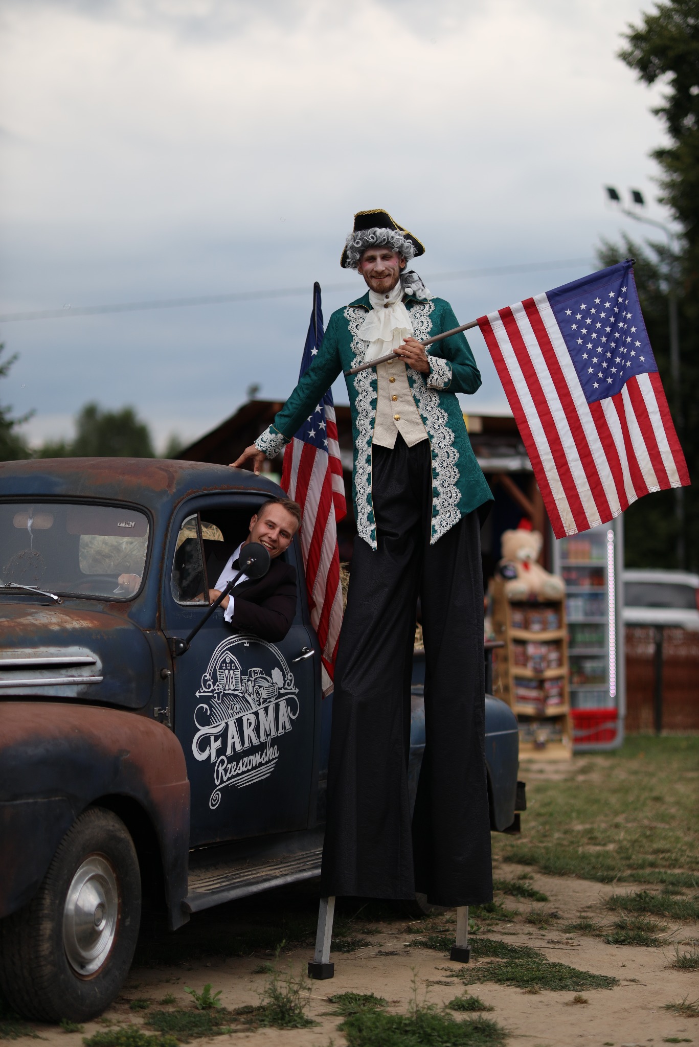 Dwóch mężczyzn: jeden na szczudłach w kostiumie z epoki, drugi w aucie z flagą USA. Nietypowa atrakcja eventowa, oryginalny sposób na urozmaicenie imprezy.