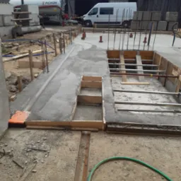 Wylany beton w szalunkach z widocznym zbrojeniem, na budowie. W tle betoniarka i biały bus. Widoczne przygotowania do dalszych prac budowlanych.