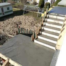 Wylany beton na tarasie z przygotowanymi schodami, widoczne drewniane szalunki i zbrojenia. W tle budynki mieszkalne i plac zabaw, ujęcie z góry.