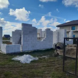 Wznoszenie ścian domu z białych bloczków. Widoczna betoniarka i sterta zaprawy. W tle budynek w trakcie budowy, błękitne niebo z chmurami.