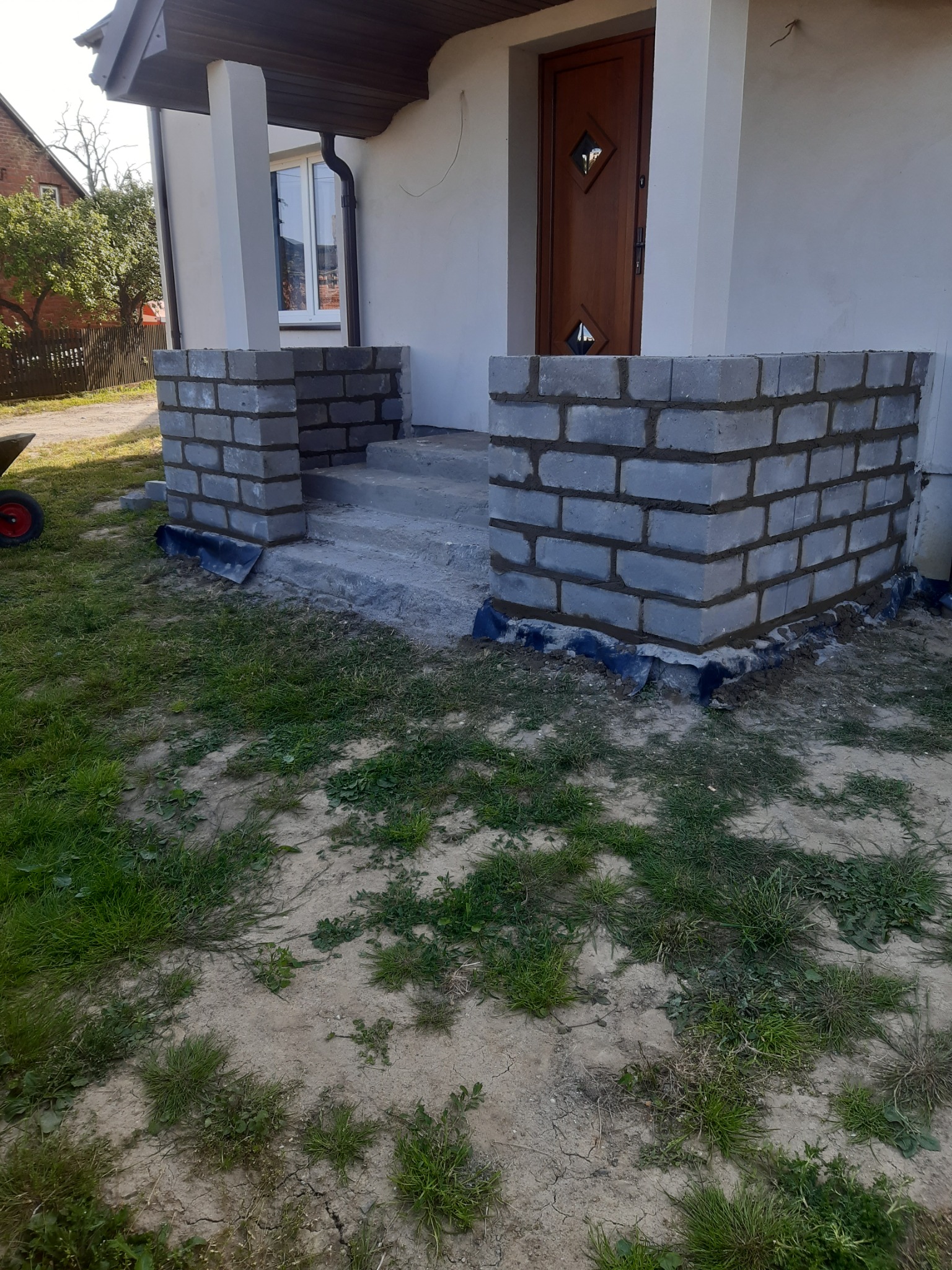 Wznoszenie muru z szarej cegły wokół wejścia do domu, widoczne fragmenty izolacji. Świeże spoiny, trawa i ziemia w tle. Częściowo zadaszone wejście.