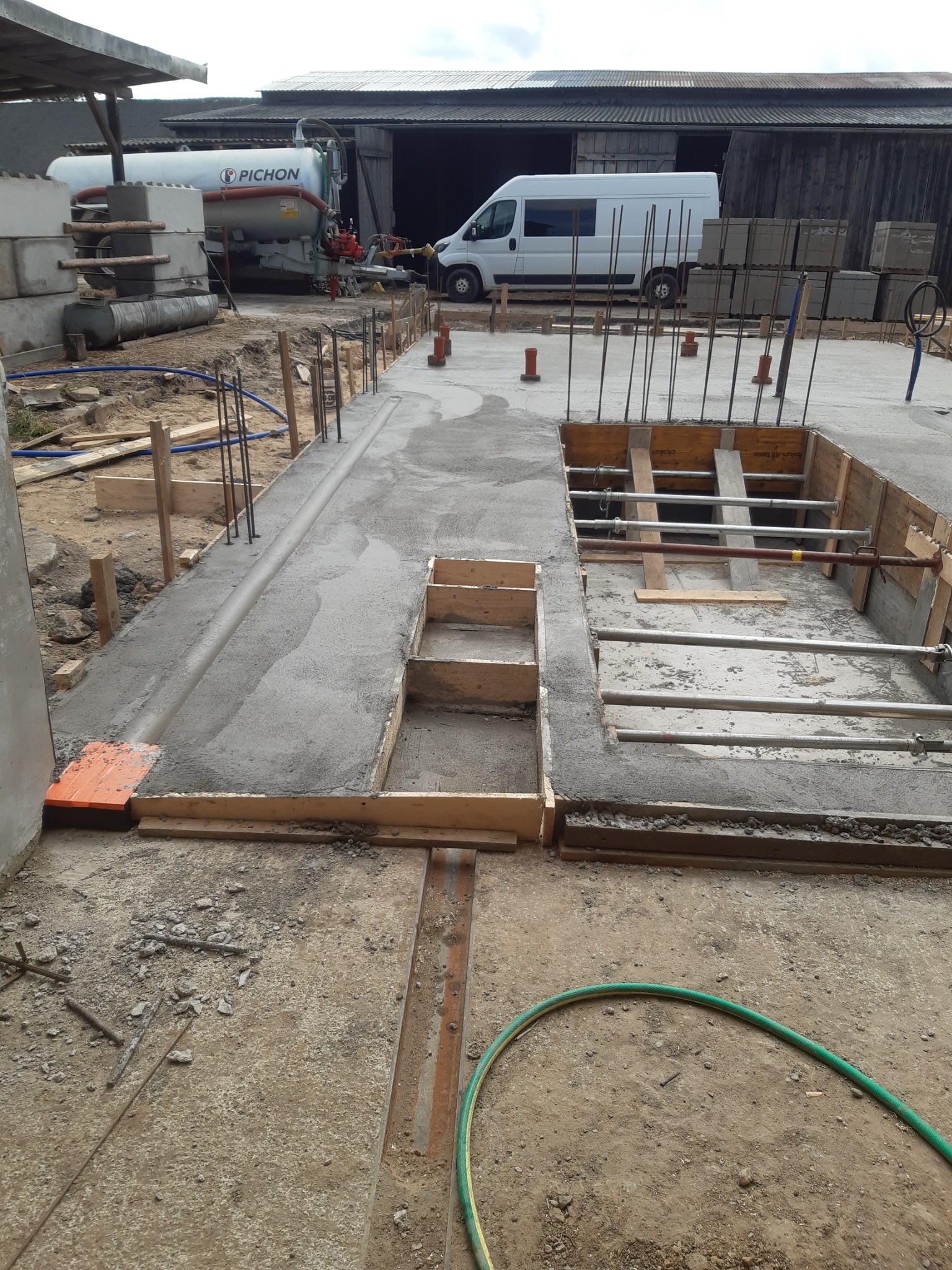 Wylany beton w szalunkach z widocznym zbrojeniem, na budowie. W tle betoniarka i biały bus. Widoczne przygotowania do dalszych prac budowlanych.