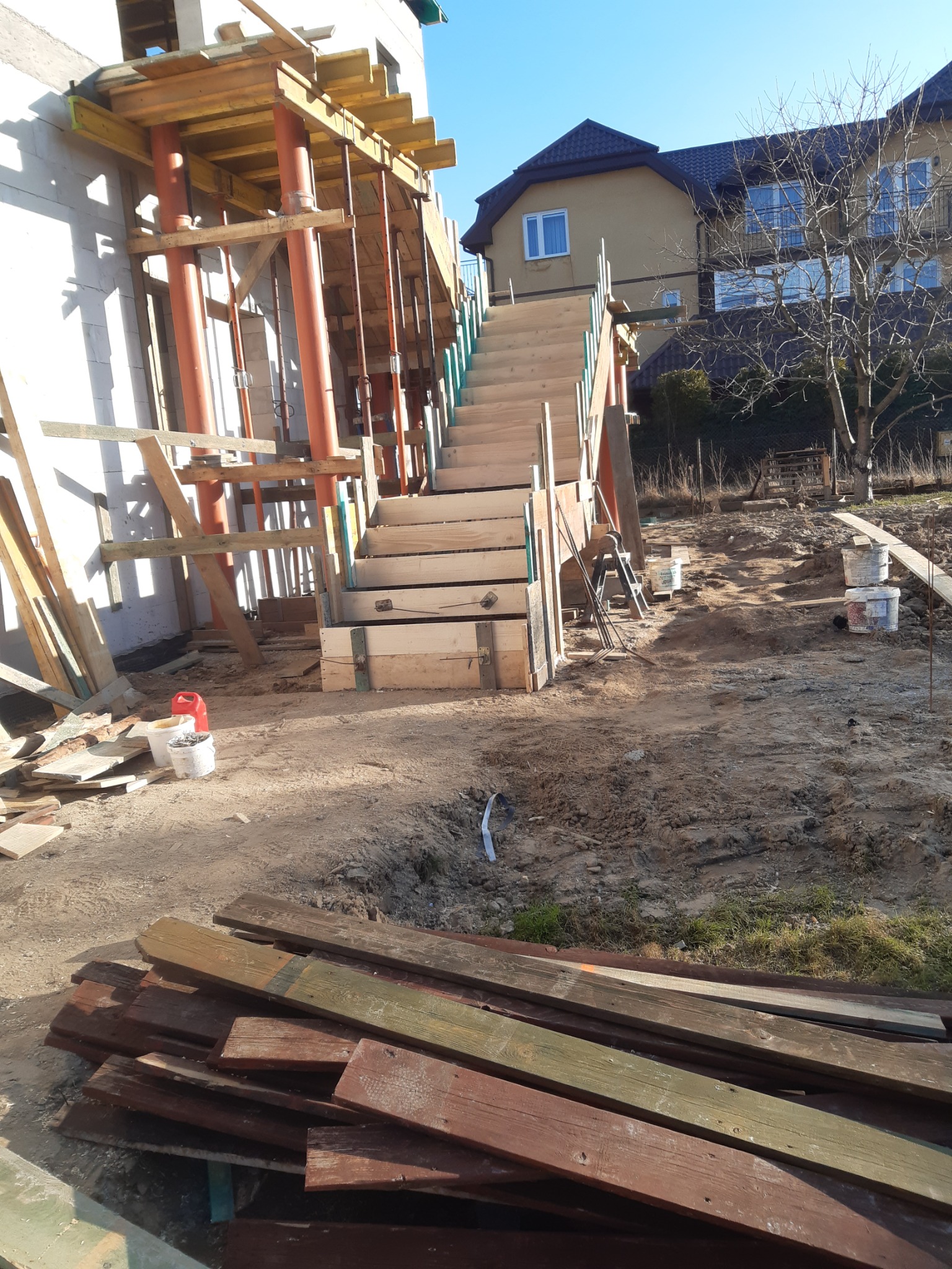 Budowa schodów zewnętrznych z drewnianym szalunkiem przy domu w trakcie budowy. Widoczne podpory, ziemia i deski na pierwszym planie. Słoneczny dzień.