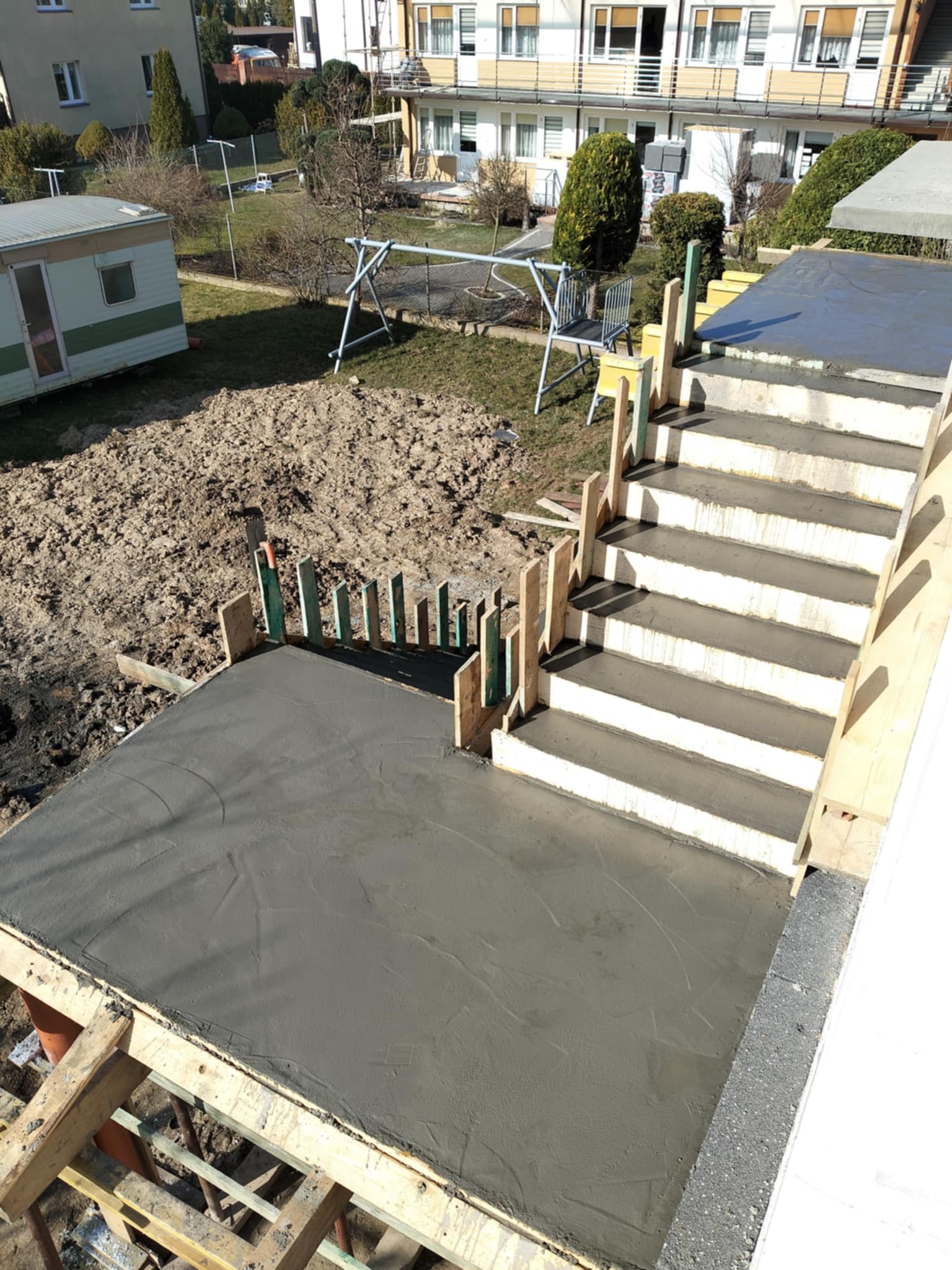Wylany beton na tarasie z przygotowanymi schodami, widoczne drewniane szalunki i zbrojenia. W tle budynki mieszkalne i plac zabaw, ujęcie z góry.