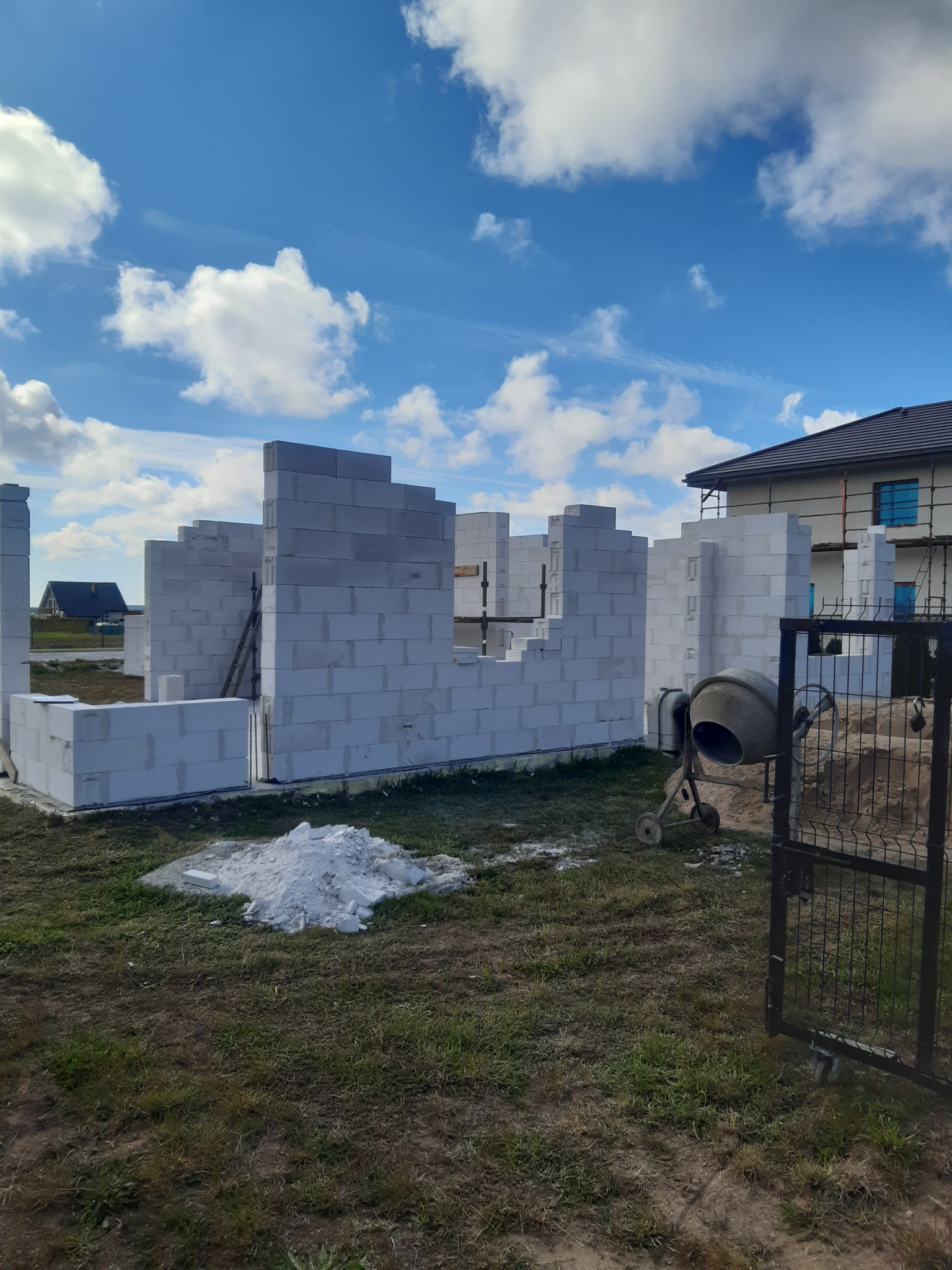 Wznoszenie ścian domu z białych bloczków. Widoczna betoniarka i sterta zaprawy. W tle budynek w trakcie budowy, błękitne niebo z chmurami.