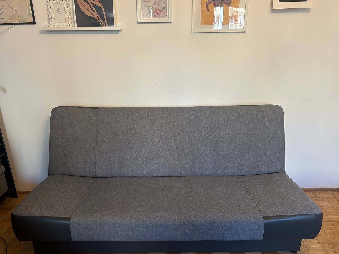 Szara, rozłożona kanapa z ciemnymi bokami na parkiecie, z obrazkami na ścianie w tle. Widok z przodu, proste ujęcie, dzienne światło. Minimalistyczny design.