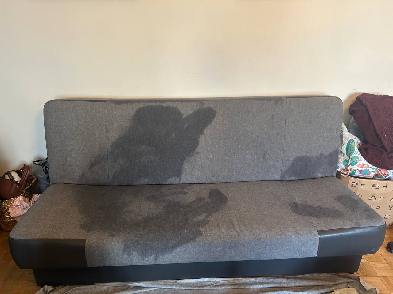 Szara, tapicerowana kanapa z wyraźnymi plamami po zalaniu, ukazująca potrzebę czyszczenia. Widoczne torby i karton z boku, podkreślające domowe otoczenie.