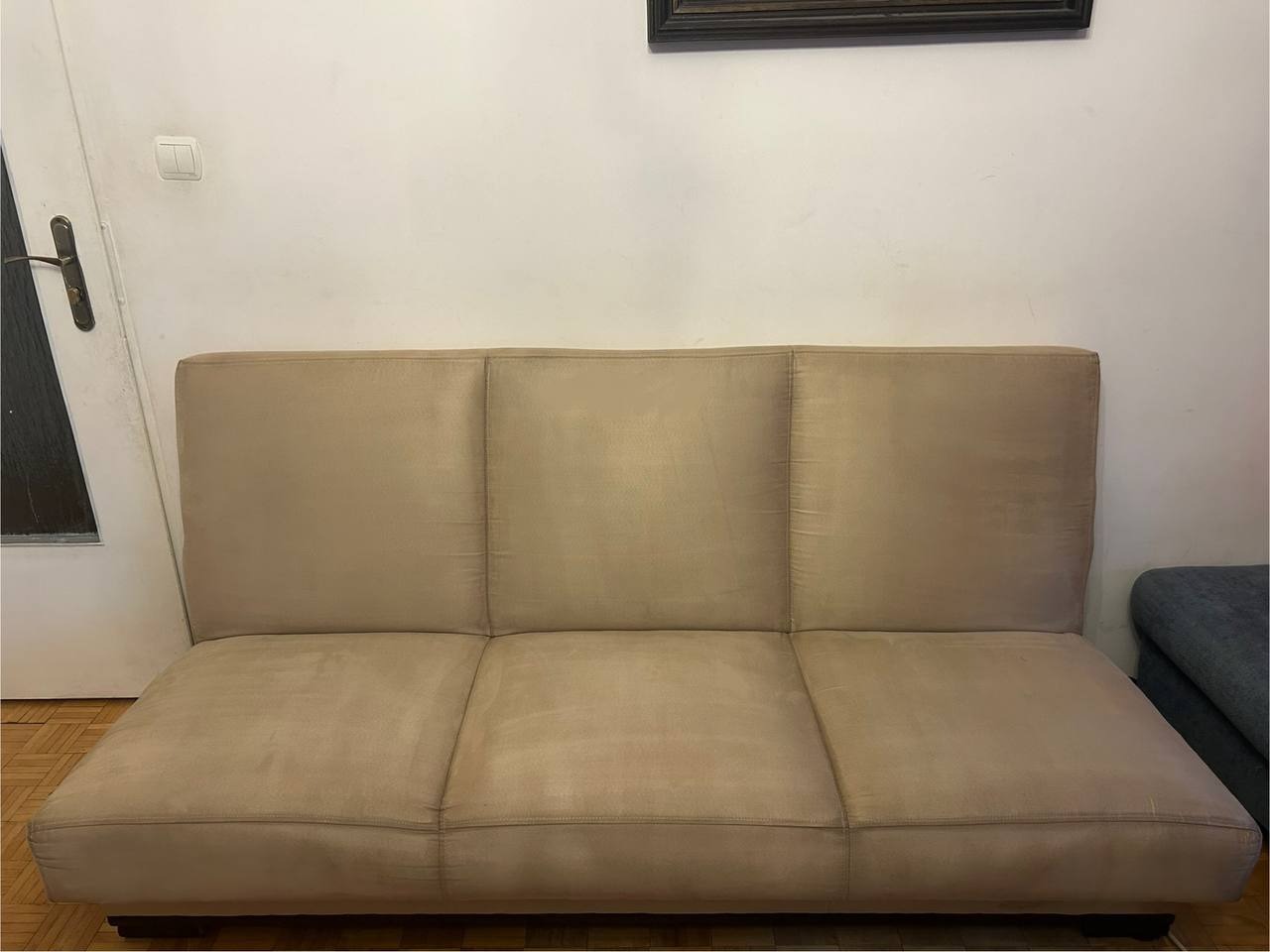 Beżowa, rozłożona sofa z wyraźnymi podziałami na trzy segmenty, ustawiona na drewnianej podłodze, na tle białej ściany z obrazem i drzwiami po lewej.