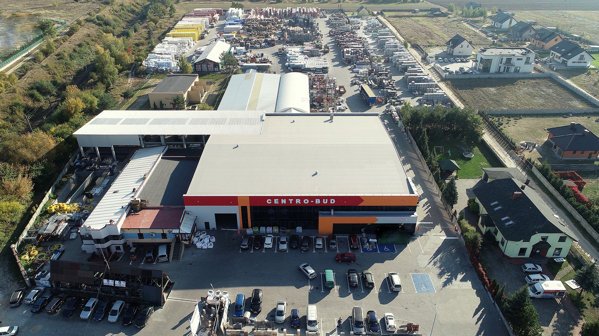 Widok z drona na rozległy skład budowlany Centro-Bud z magazynami, placem składowym pełnym materiałów i parkingiem z samochodami osobowymi. Otoczenie to zabudowa jednorodzinna.