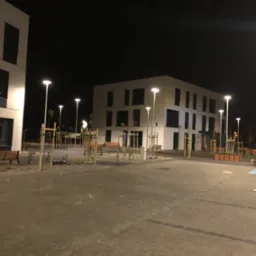 Nocny widok na plac z kostki brukowej, oświetlony latarniami, z nowo posadzonymi drzewami i budynkami w tle. Minimalistyczny styl architektury.