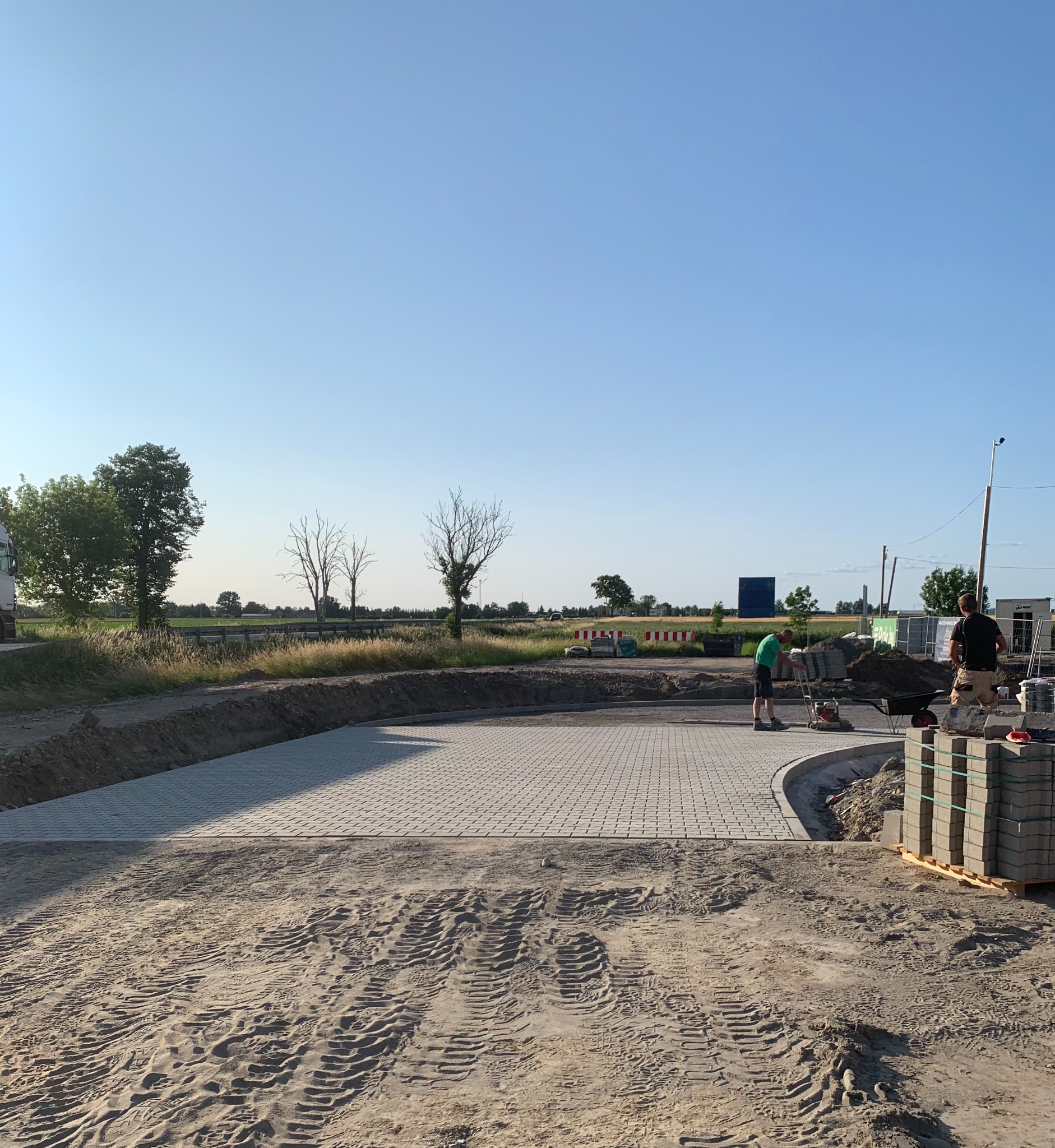 Układanie szarej kostki brukowej na przygotowanym podłożu. Widoczni dwaj pracownicy oraz maszyna do ubijania. Na pierwszym planie ślady opon na piasku.