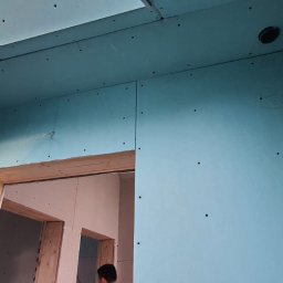 BK Plus - Montaż płyt gipsowo-kartonowych na ścianach i suficie w pomieszczeniu z drewnianą futryną drzwiową, widoczne otwory na instalacje.