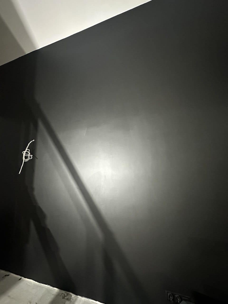 Ściana pomalowana na ciemny kolor, z widocznymi przewodami elektrycznymi. Na ścianie widoczne refleksy światła. Dolna część ściany przy podłodze jest niedokończona.