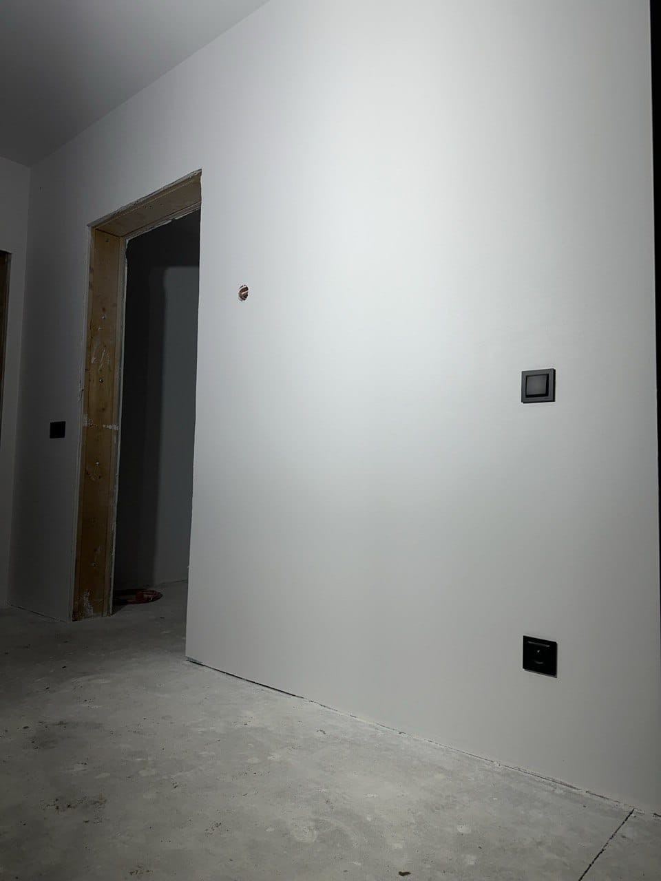 Wnętrze po gładziowaniu i malowaniu na biało, widoczne gniazdka elektryczne i surowa ościeżnica drzwiowa. Szare, betonowe podłoże. Minimalistyczny design.