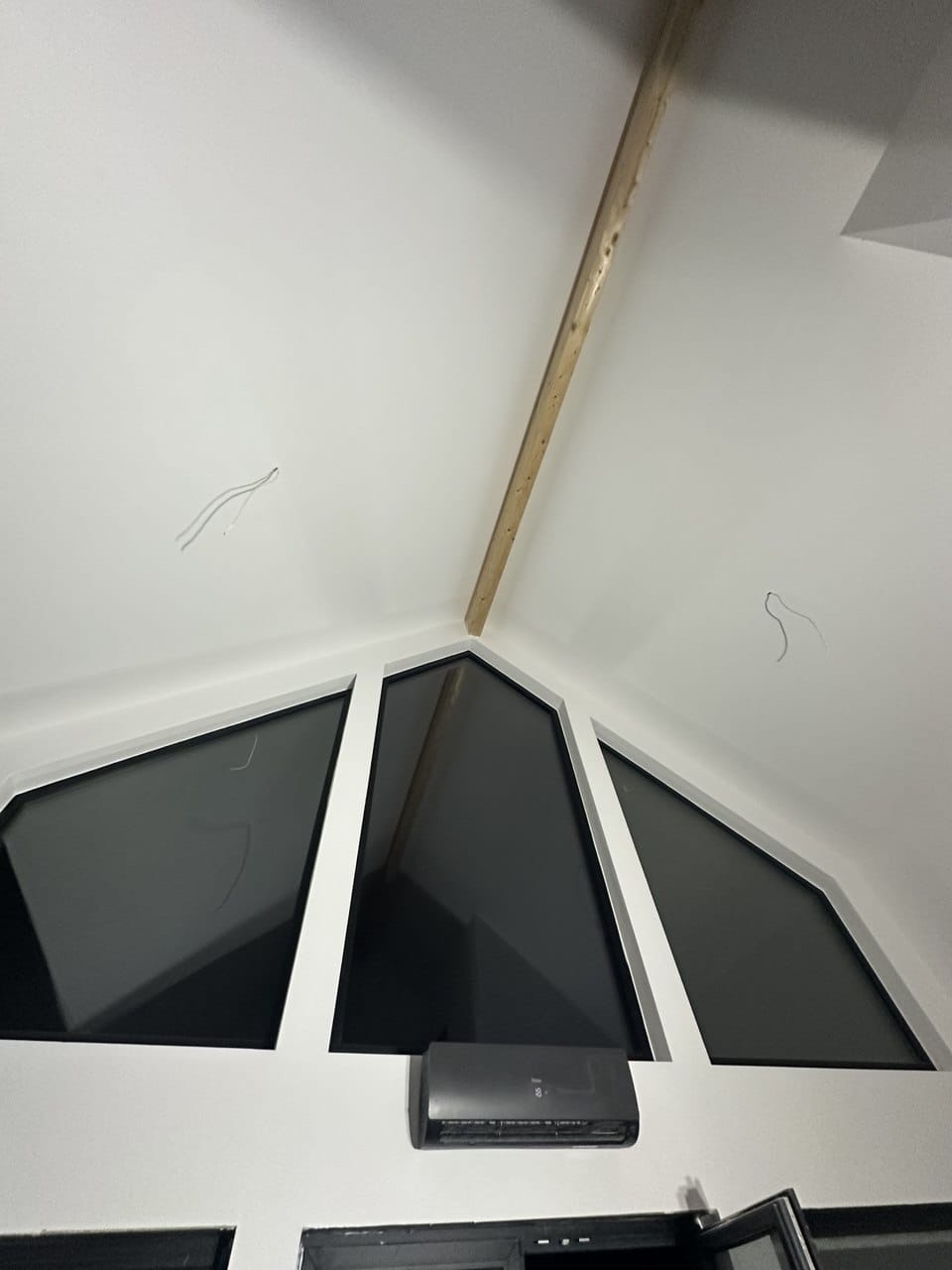 Wnętrze z oknami w kształcie trójkątów i trapezów, drewnianą belką stropową oraz zamontowanym klimatyzatorem. Widoczne wystające przewody elektryczne.