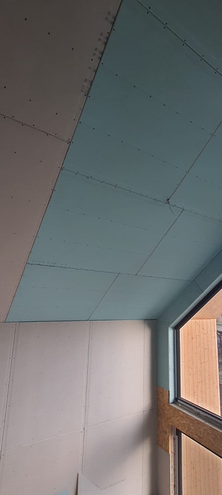 Montaż płyt gipsowo-kartonowych na suficie i ścianach, widoczne łączenia i wkręty, surowy stan wykończenia wnętrza, okno z drewnianą ramą w tle.