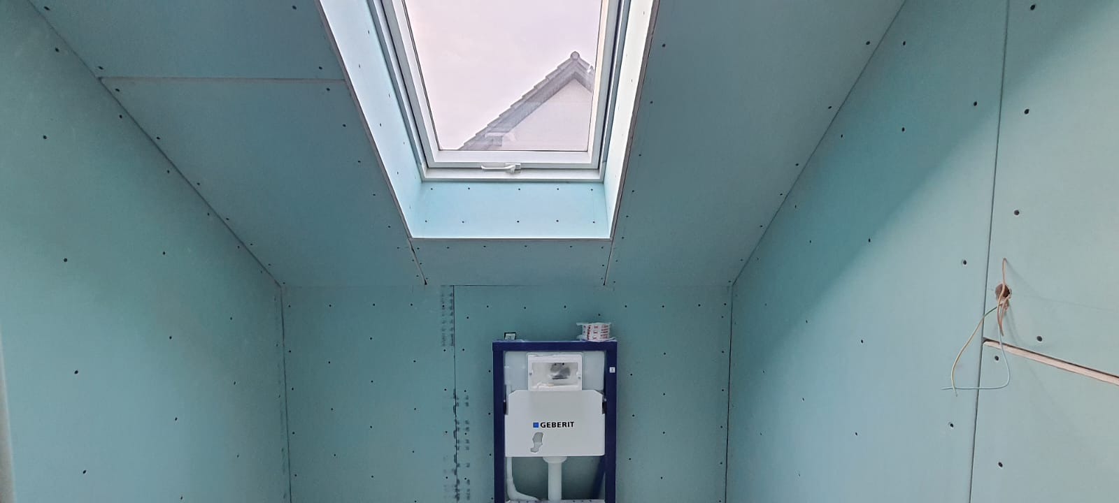 Wnętrze łazienki w trakcie remontu z zamontowanym stelażem podtynkowym Geberit i oknem dachowym, ściany pokryte płytami gipsowo-kartonowymi. Widoczne ślady po wkrętach.