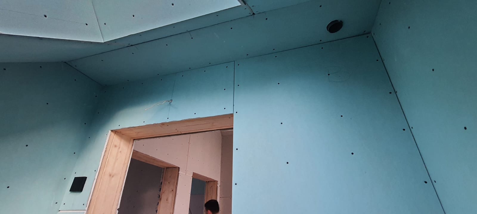 Montaż płyt gipsowo-kartonowych na ścianach i suficie w pomieszczeniu z drewnianą futryną drzwiową, widoczne otwory na instalacje.