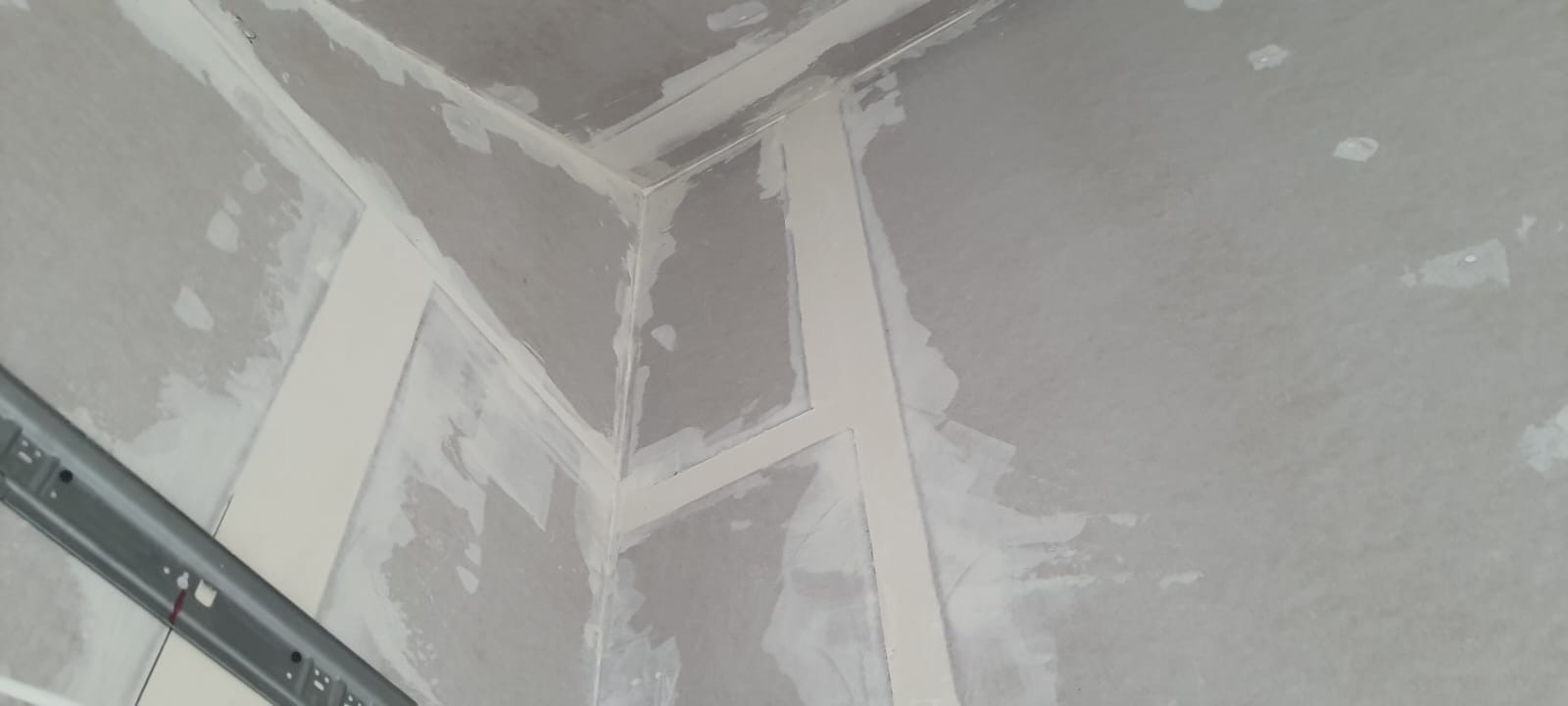 Spojenie ścian i sufitu po szpachlowaniu, widoczne białe paski taśmy wzmacniającej i szpachli. Metalowy profil przy ścianie. Ściany przygotowane do malowania.