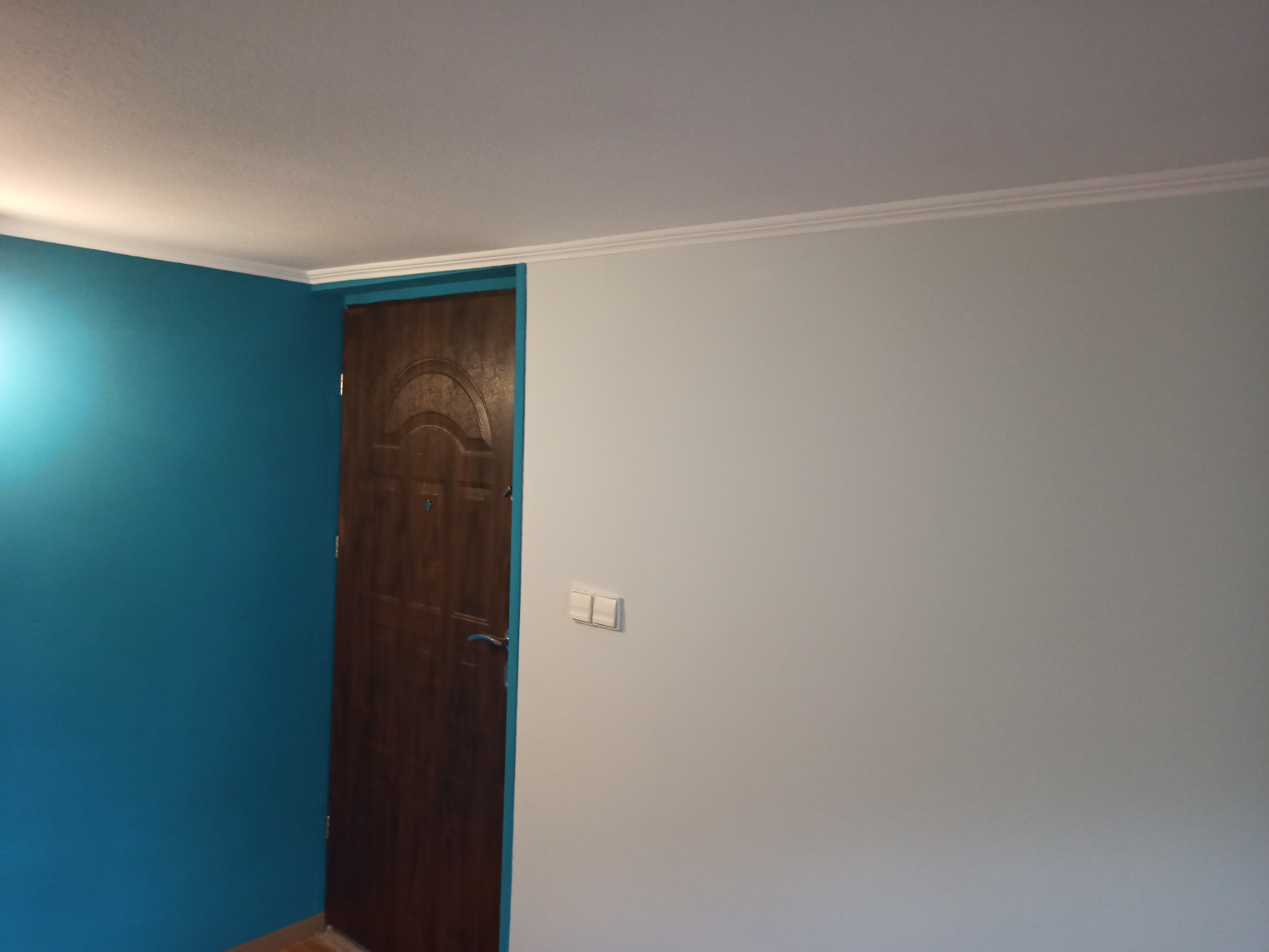 Ściana pomalowana na dwa kolory - turkusowy i szary, z brązowymi drzwiami i białą listwą przysufitową. Widoczny włącznik światła. Świeżo po malowaniu.