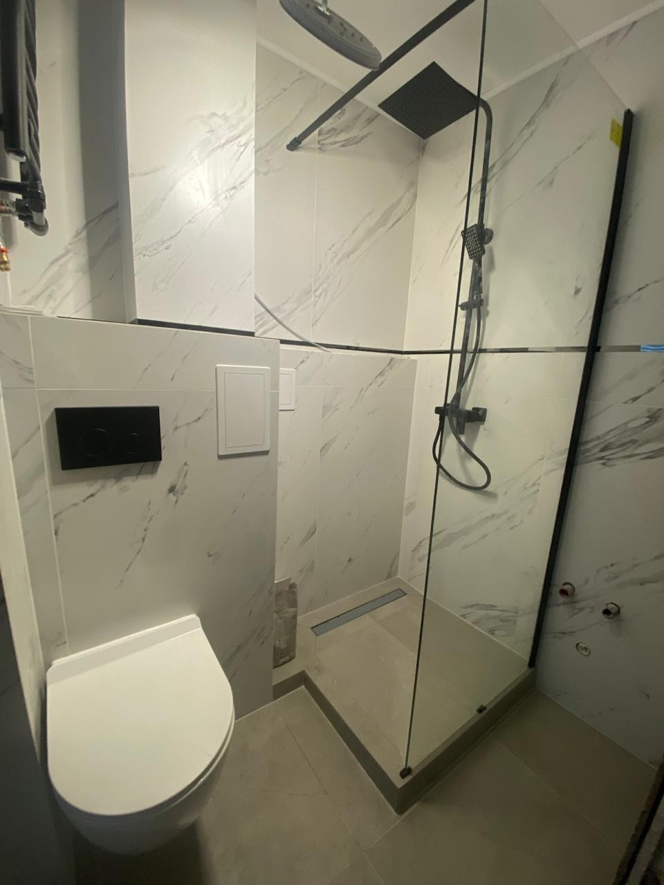 Nowoczesna łazienka z kabiną prysznicową typu walk-in, wykończona płytkami imitującymi marmur. Widoczna podwieszana toaleta i czarne elementy armatury.