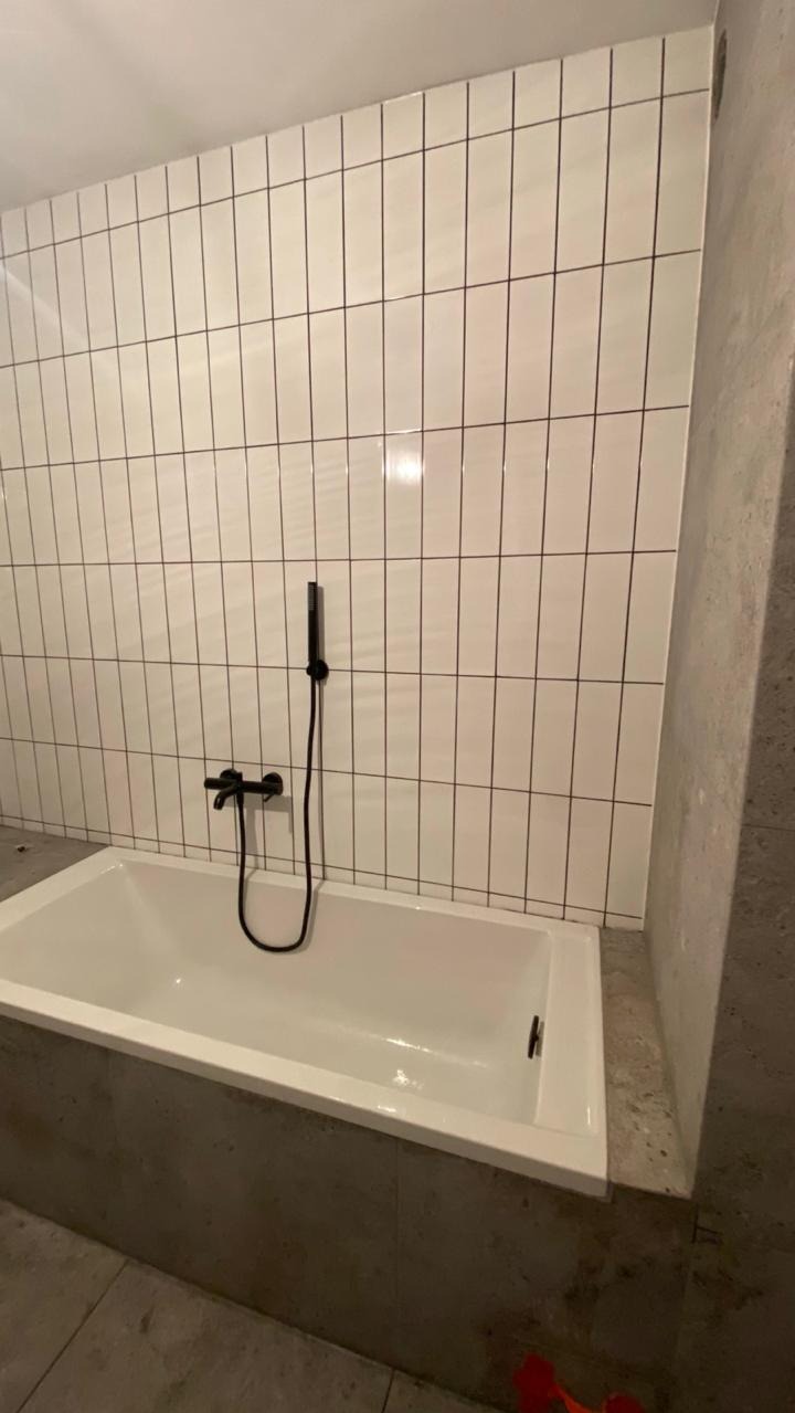 Biała wanna z czarną armaturą na tle ściany wyłożonej białymi, pionowymi płytkami z ciemną fugą. Minimalistyczny design łazienki po remoncie.