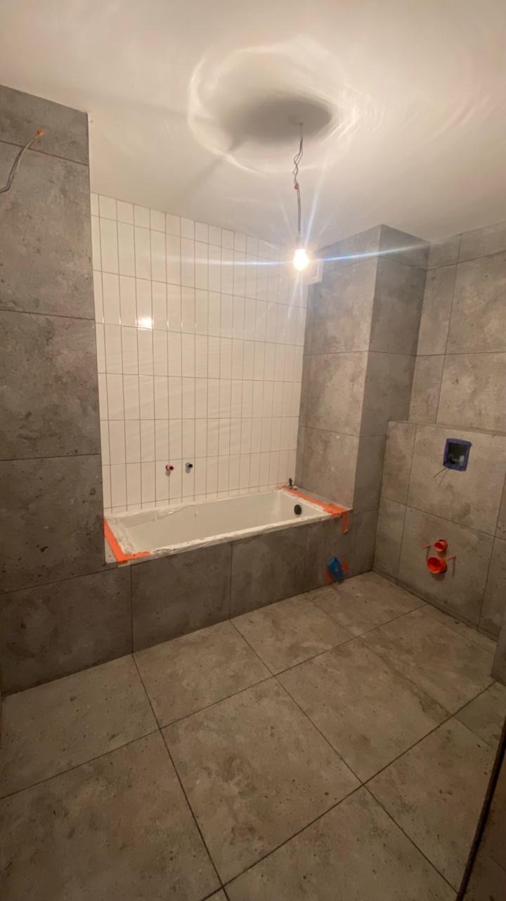 Remont łazienki w trakcie: białe płytki nad wanną, szare płytki na ścianach i podłodze. Widoczne instalacje i zabezpieczenia. Oświetlenie sufitowe.