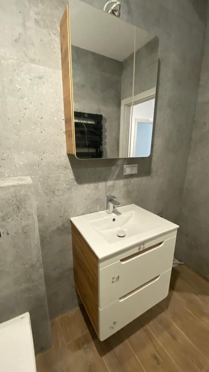 Nowoczesna łazienka z umywalką na szafce z drewnianymi bokami, lustrem w drewnianej ramie i szarymi płytkami imitującymi beton. Widoczny grzejnik drabinkowy.